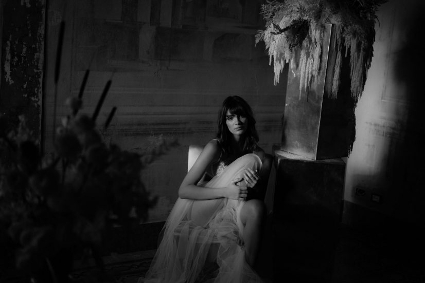 Black and white portrait of a seated bride beside flowers in a shadowed historic room